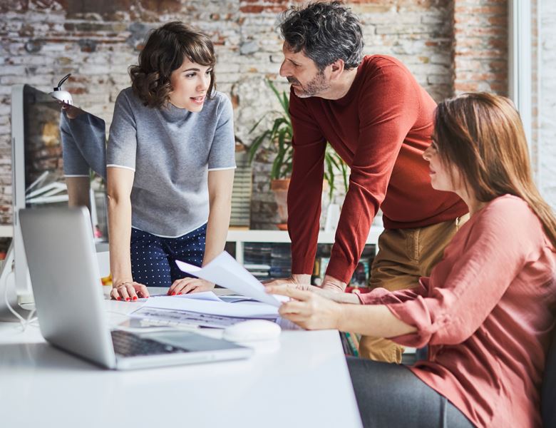 Co-workers leaning over a desk and computer while discussing something.