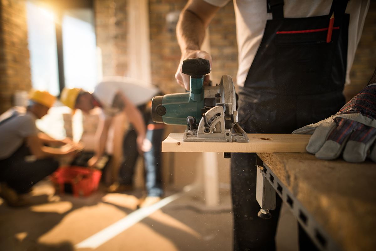 Someone using a saw to cut a board with two other workers in the background.