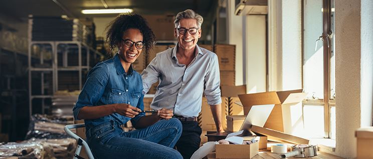 A man and a woman in a warehouse, smiling and boxing up items.