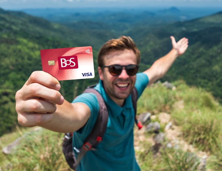 A man holding a BOS debit card while standing on the top of a green mountain.