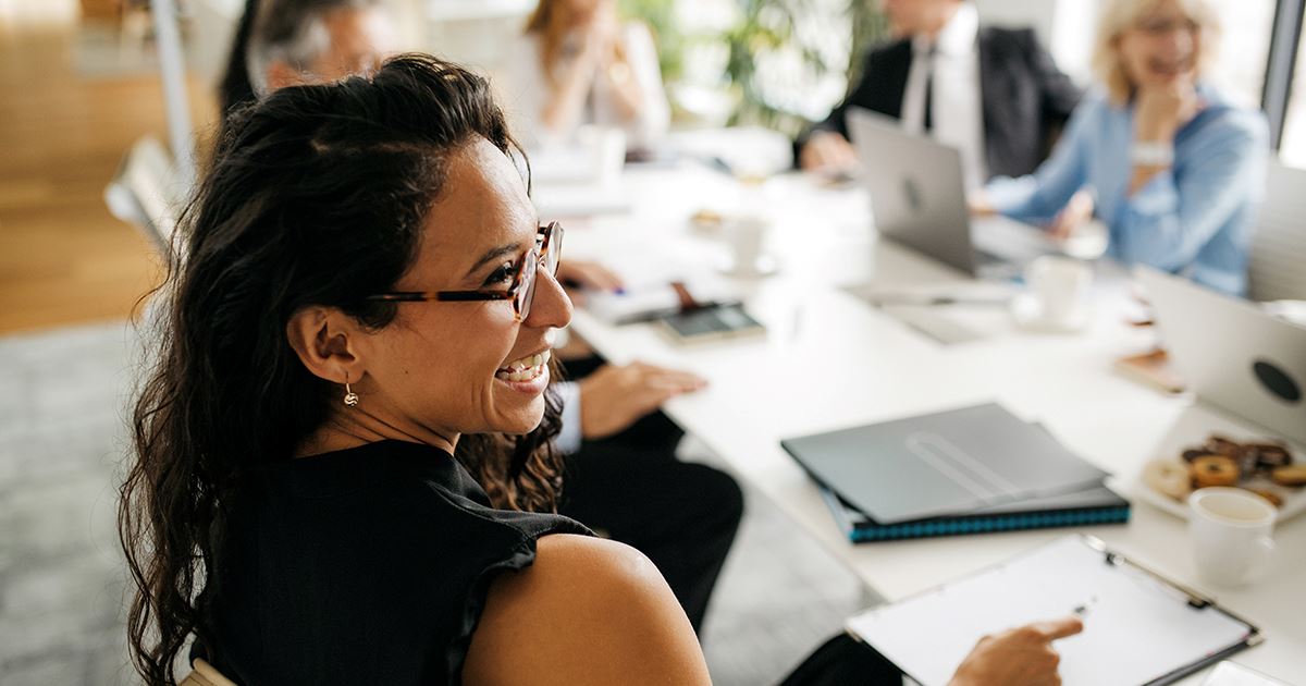 A smiling business woman sitting with cowokers around a conference room table with laptops and paperwork.