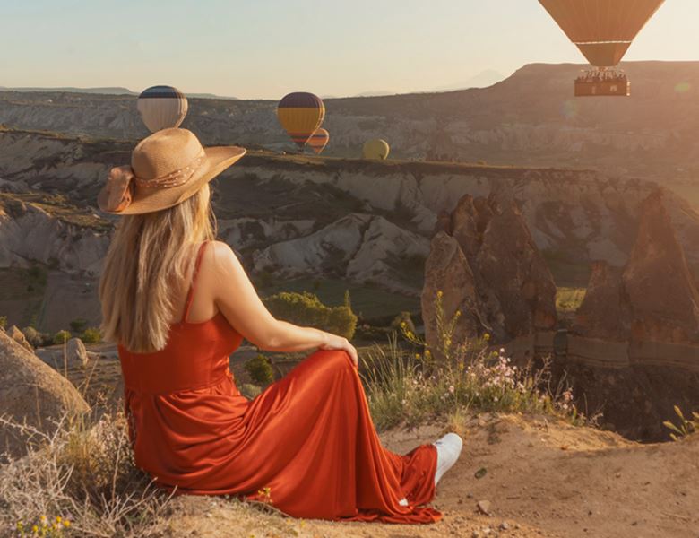 A woman sitting on a mountainside watching hot air balloons fly by.