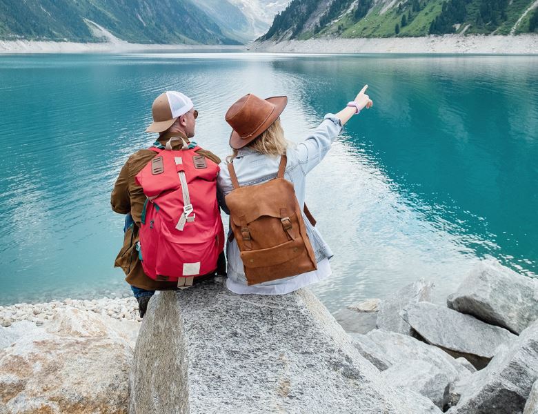 A man and woman sitting on the edge of a lake with mountains in the background.  The woman is pointing at something.
