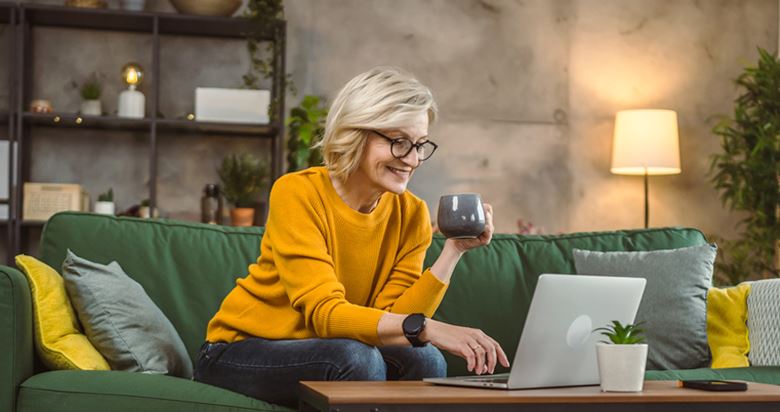 A woman sitting on the couch looking at her laptop and holding a coffee cup.