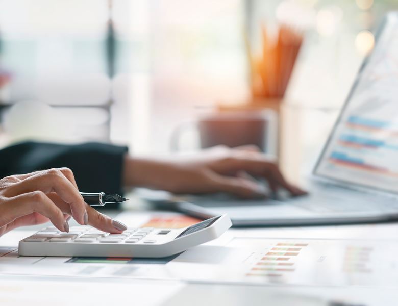 A picture showing a woman's hands typing on a calculator and laptop with graphs on the desk.