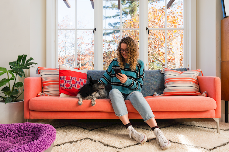 A woman sitting on a couch next to a dog while checking her mobile phone.